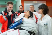 Dr. Haase (Fabian Harloff, l.) hat Schwester Adriana (Christina Geiße, liegend) versprochen, weder Dr. Jonas (Gerit Kling, r.) noch sonst jemandem ihr Geheimnis zu verraten. Zwei weitere Sanitäter (Komparsen) helfen beim Transport.