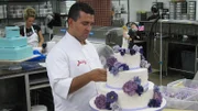 Buddy adds flowers to back of same sex cake, and Krista decorates in background.