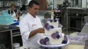 Buddy adds flowers to back of same sex cake, and Krista decorates in background.