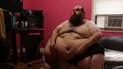 Allen is sitting on a bed, shirtless.