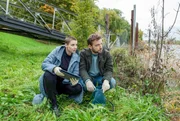 Nina (Julia E. Lenska, l.) und Gregor (Jonas Minthe, r.) sitzen im Gras vor dem Bootssteg und durchsuchen mit ernsten Mienen die Wertsachen von Tessa.