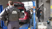 Ein CBP-Beamter arbeitet an der Grenze von San Ysidro daran, Pakete mit mutmaßlichen Betäubungsmitteln aus dem Fahrzeug eines Verdächtigen zu bergen, während ein Kameramann filmt und ein anderer CBP-Beamter zusieht.