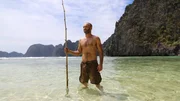 Ed Stafford standing, holding a bamboo rod with the sea and cliffs behind him.