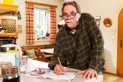 Stadlbauer (Andreas Geiß) löst ein Kreuzworträtsel hinter Monikas Rücken.