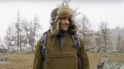 Ed Stafford close-up wearing fur hat with snowy trees and rock in background.