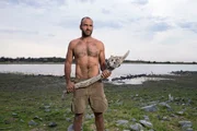 Ed Stafford holding a tool he has made in front of watering hole landscape, Namibia.
