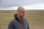Ed Stafford l&auml;chelt in einer grauen Jacke und blickt in einer ruhigen Landschaft in der Mongolei nach rechts.