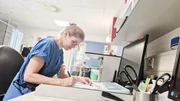 E7 - Female physician with blue scrubs writing a report while sitting at a desk with a computer in front of her. To the right there are scissors in a cup