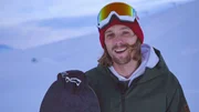 Eine Person steht in einer verschneiten Umgebung und h&auml;lt ein Snowboard, gekleidet in eine gr&uuml;ne Jacke, einen wei&szlig;en Hoodie, eine rote M&uuml;tze und Skibrille.