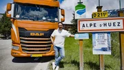 Ep7 - Sebastian in front of the truck (truck on the left of the frame,) left hand leaning on a Alpe d’Huez sign, right thumb on his side, smiling, looking straight to the camera, total