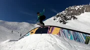 Ein Snowboarder f&uuml;hrt einen Trick aus, w&auml;hrend er auf einer farbenfrohen Rail im Schneepark einer Bergkulisse gleitet.