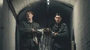 SOE-Agent wird in einem Lager durch einen Flur getragen.