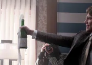 Eine Person in einem Anzug h&auml;lt eine Flasche Champagner, aus der reichlich Schaum sprudelt, w&auml;hrend jemand im Hintergrund diesen Moment fotografiert.