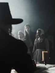 A young man stands guard over two accused witches, both of whom are facing the council in front of them.