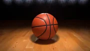 A basketball on a wood floor beneath bright arena lights