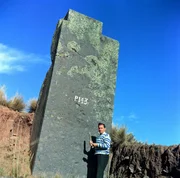 Ein Mann steht neben einem gro&szlig;en Steinmonolithen mit der Markierung &bdquo;P143&ldquo; in einer offenen Landschaft.