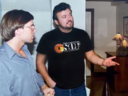 Zwei M&auml;nner unterhalten sich in einem Innenraum, wobei einer mit der Hand gestikuliert und der andere ihm gegen&uuml;bersteht.