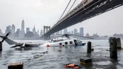 Ein medizinischer Evakuierungshubschrauber mit der Aufschrift &bdquo;MEDIVAC&ldquo; liegt teilweise im Wasser versunken nahe einer Br&uuml;cke mit der Skyline von New York City im Hintergrund; Tr&uuml;mmer treiben in der Umgebung.