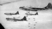 Vier Militärflugzeuge fliegen in Formation, jedes mit einem „X“-Symbol am Heck gekennzeichnet. Bomben fallen aus den Flugzeugen und zeigen einen Moment der Luftkriegsführung, vermutlich aus einem historischen Konflikt.