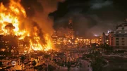 Ein gewaltiges Feuer verschlingt mehrere Geb&auml;ude in einem st&auml;dtischen Gebiet bei Nacht &ndash; lodernde Flammen und dichter Rauch steigen in den Himmel. Im Vordergrund sind einige Personen zu sehen, die m&ouml;glicherweise fliehen oder das Geschehen beobachten.