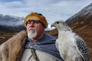 Ein Reenactor-Wikinger und ein Gerfalke (Gyrfalcon) in der verschneiten Landschaft des Alladale Naturschutzgebietes in Schottland.