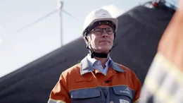 Uwe Braun auf seinem Werksgel&auml;nde vor einem Berg mit Eisenerz und werkseigenem Windrad.