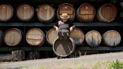 Ein Mann mit Strohhut lehnt entspannt an einem großen Weinfass, umgeben von weiteren Fässern. Die Szenerie strahlt eine ruhige, ländliche Atmosphäre aus und spiegelt den traditionellen Charme der südsteirischen Weinstraße wider.