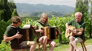 Drei Personen spielen Musikinstrumente im Freien, umgeben von Grün und einer Berglandschaft. Zwei spielen Akkordeon, die dritte spielt Gitarre. Alle tragen grüne T-Shirts und sitzen auf Holzstühlen. Die Atmosphäre ist entspannt und fröhlich.