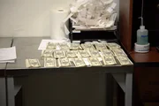 Stapel von Falschgeld werden auf einen Inspektionstisch gelegt, nachdem sie in mehreren handgefertigten Bilderrahmen im Gep&auml;ck eines Passagiers in Atlanta, Georgia, entdeckt wurden (National Geographic for Disney).