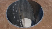 Das Bild zeigt eine große kreisförmige Ausgrabung im Boden, die mit Betonwänden ausgekleidet ist. Das Bild zeigt eine große kreisförmige Ausgrabung im Boden, die mit Betonwänden ausgekleidet ist.