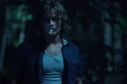 A young man smokes a cigarette in the woods.