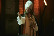 Der Bischof von Arl (Max Giermann) lässt (fast) nichts anbrennen und antwortet einer Hexe auf dem Scheiterhaufen im Rahmen eines Musicals.
