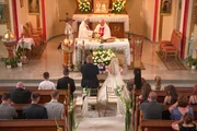 Photograph from the church. The priests are standing behind the altar, the bridal couple is in front of them. The wedding guest are kneeing on the church benches.