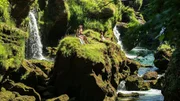 Menschen sitzen auf einem sonnenbeschienenen, grasbewachsenen Felsen neben kleinen Wasserf&auml;llen &uuml;ber einem klaren Fluss.