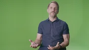 Eine Person sitzt vor einem einfarbigen gr&uuml;nen Hintergrund und tr&auml;gt ein kurz&auml;rmeliges, dunkelblaues Hemd mit Kn&ouml;pfen. Die H&auml;nde sind ge&ouml;ffnet und gestikulieren, was auf einen erkl&auml;renden oder Gespr&auml;chsmoment in einem Studio hinweist.