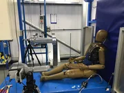 Ein Crashtest-Dummy sitzt auf einer blauen Plattform in einer Prüfeinrichtung und lehnt an einer gepolsterten vertikalen Fläche. Der Dummy ist mit Sensoren und Verkabelungen ausgestattet, und eine blaue Weste bedeckt den Oberkörper. Um den Dummy herum befinden sich verschiedene Prüfgeräte, darunter Roboterarme, Kabel und Strukturrahmen, was auf eine kontrollierte Umgebung für Fahrzeugsicherheitsprüfungen hinweist.