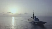 Ein modernes Kriegsschiff f&auml;hrt &uuml;ber ruhige Gew&auml;sser bei Sonnenaufgang oder Sonnenuntergang. Das Schiff hat ein schlankes, kantiges Design mit einem hohen Mast und Radarausr&uuml;stung, die sich als Silhouette vor dem hellen Horizont abzeichnet.