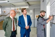 Nachdem Hausmeister Winkler (Steffen Wolf, r.) die neuen Schilder montiert, sind Markus Herrmann (Peter Ketnath, M.) und Polizeidirektor Achtziger (Alexander Duda, l.) überzeugt, dass sich nun niemand mehr verlaufen wird.
