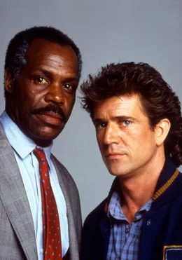 Die Cops Martin Riggs (Mel Gibson, r.) und Roger Murtaugh (Danny Glover) finden ein Auto, bei dem der Kofferraum mit Krugerrand-Goldm&uuml;nzen gef&uuml;llt ist. Woher stammen die vielen M&uuml;nzen?