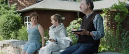Mutter Vera (Eva L&ouml;bau), Tom (Theo Kretschmer) und Opa Horst (Michael Wittenborn) sitzen vor dem Haus auf der Steinmauer und essen Apfelkuchen. Tom und Vera verabschieden sich voneinander.