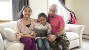 Marlow Williamson sitzt mit seinen T&ouml;chtern Helena Williamson und Isabella Williamson auf dem Sofa. Die J&uuml;ngste h&auml;lt ein iPad, das Marlow beim Erkl&auml;ren anschaut; die beiden M&auml;dchen schauen in die Kamera und l&auml;cheln.