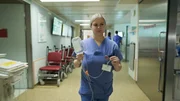 Portrait of a young blonde female nurse walking on the aisle carrying an IV bag with one hand and holding its tube with the other. To her back we see several dark red wheelchairs lined up on the wall. Also two colleagues are in the background also wearing blue scrubs.