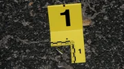Eine Patronenh&uuml;lse liegt auf einer Asphaltoberfl&auml;che neben einem gelben Beweismarker mit der Aufschrift &bdquo;1&ldquo;, was auf ihre Dokumentation am Tatort hinweist.