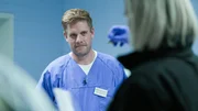 Ep6 - A portrait of Dr Zimmermann smiling lightly while listening to the female paramedic with the grey bob, who is showing something in her covered hand with blue gloves. The paramedic is in the front but blurry and is also wearing a black jacket.