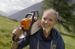 Holzbildhauerin Nora Engels l&auml;uft mit Motors&auml;ge auf der Schulter einen Berghang im Oberengadin hinauf.