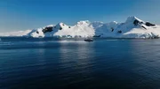 Das Bild zeigt ein Kreuzfahrtschiff, das durch antarktische Gew&auml;sser f&auml;hrt; im Hintergrund sind schneebedeckte Berge zu sehen.