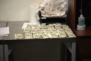 Stapel von Falschgeld werden auf einen Inspektionstisch gelegt, nachdem sie in mehreren handgefertigten Bilderrahmen im Gep&auml;ck eines Passagiers in Atlanta, Georgia, entdeckt wurden (National Geographic for Disney).