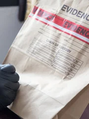 Forensic hand in black glove writing on evidence bag and sealing it with red tape at a crime scene. Forensic hand in black glove writing on evidence bag and sealing it with red tape at a crime scene.