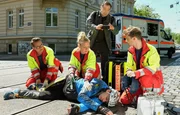 W&auml;hrend der Notarzt (Markus Neumann, li.) und seine beiden Kollegen (Jascha Rust (re.) und Sarah Zeising (mi)) den verletzten Fahrradfahrer (Tim Haberland, liegend) versorgen, filmt ein Gaffer (Albrecht Ganskopf) die ganze Zeit mit.