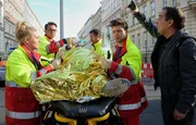 W&auml;hrend der Notarzt (Markus Neumann, 2.v.li.) und seine Kollegen (Jascha Rust (2.v.re.) und Sarah Zeising (li.), Komparse, 3.v.re.) den verletzten Fahrradfahrer (Tim Haberland, liegend) versorgen, filmt ein Gaffer (Albrecht Ganskopf) die ganze Zeit mit und steht den Rettungskr&auml;ften st&auml;ndig im Weg.